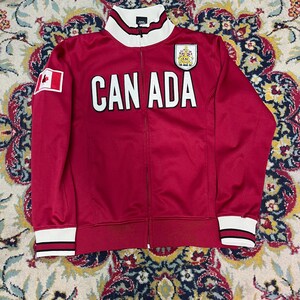 Op de afbeelding: Een rode trainingsjas met ritssluiting en het woord "CANADA" in grote witte letters. Het heeft een Canadese vlag patch, een wapen en wit en zwart gestreepte manchetten en kraag. De jas is gemaakt van een glad, glanzend materiaal.