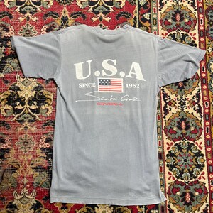 May include: A pale blue t-shirt featuring the text "U.S.A" above an American flag graphic. Below the flag, the text reads "Santa Cruz O'Neill" and "Since 1952". The shirt is made of a soft material.