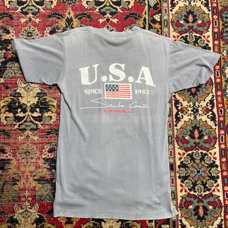May include: A pale blue t-shirt featuring the text "U.S.A" above an American flag graphic. Below the flag, the text reads "Santa Cruz O'Neill" and "Since 1952". The shirt is made of a soft material.