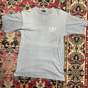 May include: A pale blue t-shirt with short sleeves. The shirt displays the text "U.S.A" with a small American flag graphic and the text "Since 1783". The shirt is made of a soft material and has a crew neck.