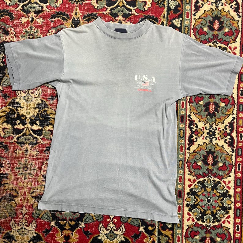 May include: A pale blue t-shirt with short sleeves. The shirt displays the text "U.S.A" with a small American flag graphic and the text "Since 1783". The shirt is made of a soft material and has a crew neck.