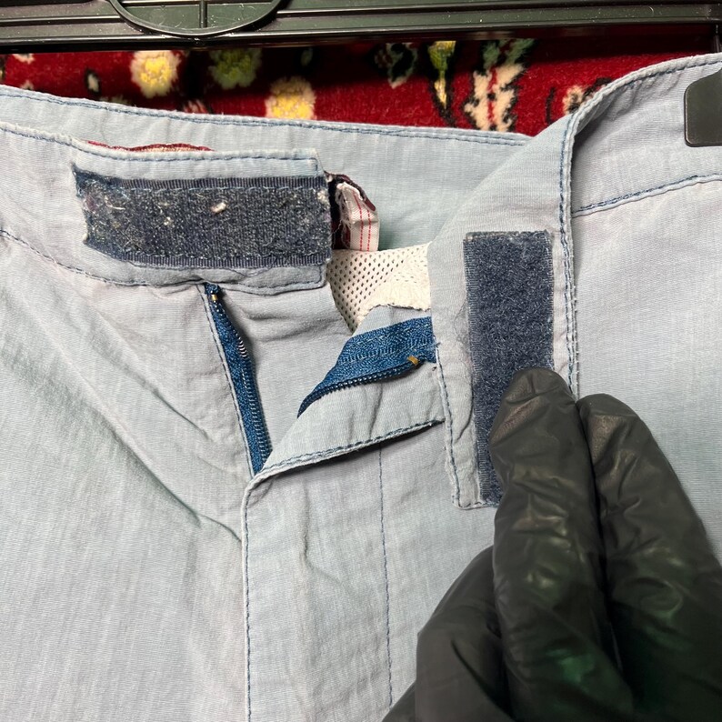 Puede incluir: Primer plano de unos pantalones cortos azul claro con cremallera y cierres de velcro. Los pantalones cortos est&aacute;n hechos de una tela ligera y tienen una cremallera azul. Los cierres de velcro son de color azul oscuro.
