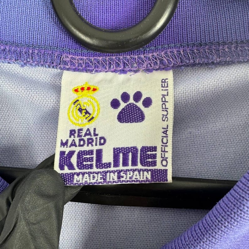 Puede incluir: Primer plano de una etiqueta de una camiseta del Real Madrid. La etiqueta es blanca con el logo del Real Madrid, una huella de pata y el logo de Kelme. El texto "MADE IN SPAIN" es visible. La camiseta es morada y azul.