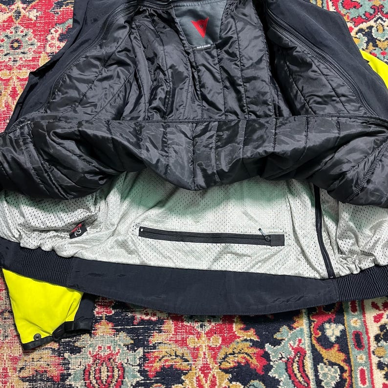 May include: Black and yellow motorcycle jacket with a quilted black lining. The jacket has a zippered pocket and a mesh interior. The Dainese logo is visible on the inside of the jacket.