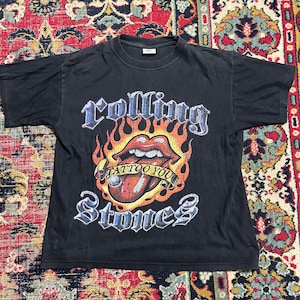 Puede incluir: Camiseta negra con el logo de los Rolling Stones, una lengua roja, llamas y las palabras "Tattoo You". El diseño incluye el nombre de la banda en una fuente gótica. Una camiseta clásica de rock and roll.