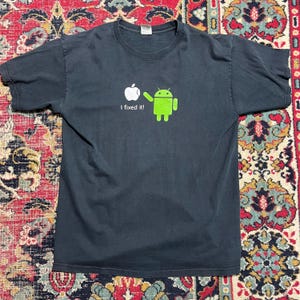 May include: Dark gray t-shirt featuring a white apple logo pointing to a green Android robot. The text "I fixed it!" is printed below the apple. The shirt is a short-sleeved crew neck.
