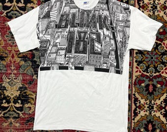 May include: White t-shirt with a black and white graphic design featuring the text "BLINK 182" in large letters. The graphic depicts a cityscape with buildings and architectural details. The shirt has short sleeves and a crew neck.
