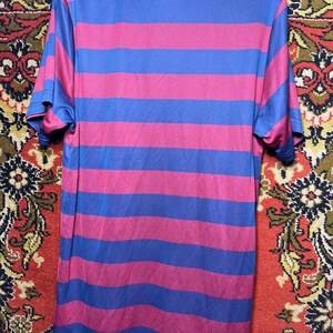 Peut inclure: Un t-shirt &agrave; manches courtes avec des rayures horizontales alternant le bleu et le magenta. Le t-shirt a un col rond avec un col gris clair. Le tissu semble &ecirc;tre un mat&eacute;riau l&eacute;ger et respirant.