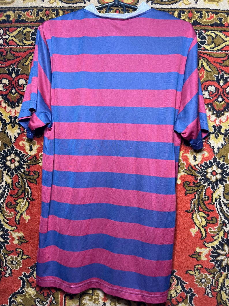Peut inclure: Un t-shirt &agrave; manches courtes avec des rayures horizontales alternant le bleu et le magenta. Le t-shirt a un col rond avec un col gris clair. Le tissu semble &ecirc;tre un mat&eacute;riau l&eacute;ger et respirant.