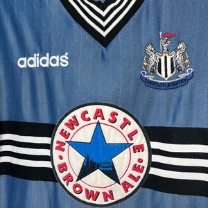 May include: A light blue Adidas soccer jersey with a V-neck collar and black and white trim. The jersey features the Newcastle United crest, the Adidas logo, and a circular patch with the text "Newcastle Brown Ale".
