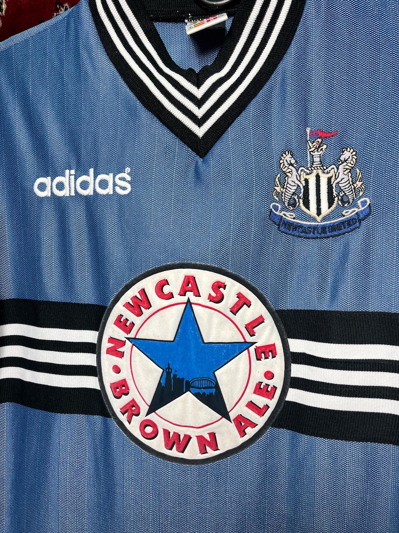 May include: A light blue Adidas soccer jersey with a V-neck collar and black and white trim. The jersey features the Newcastle United crest, the Adidas logo, and a circular patch with the text "Newcastle Brown Ale".