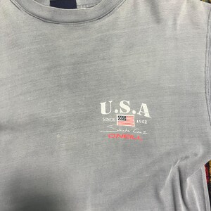 May include: A pale grey t-shirt featuring the white text "U.S.A." above a small American flag graphic, with "SINCE 1952" and "Santa Cruz O'NEILL" in red. The shirt has a crew neck and short sleeves.