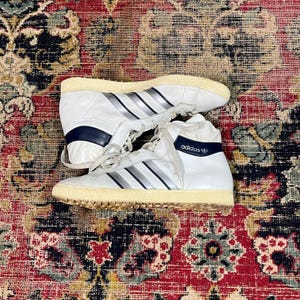 May include: Pair of vintage Adidas high-top sneakers in white leather with silver stripes. Features a cream-colored sole and navy blue accents. The Adidas logo is visible on the side. The sneakers are in good condition.