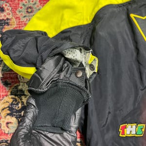 May include: Black and yellow jacket with a star design and the word "THE" in a colorful patch. The jacket has a ribbed cuff and a snap closure. The sleeve is black with yellow accents.