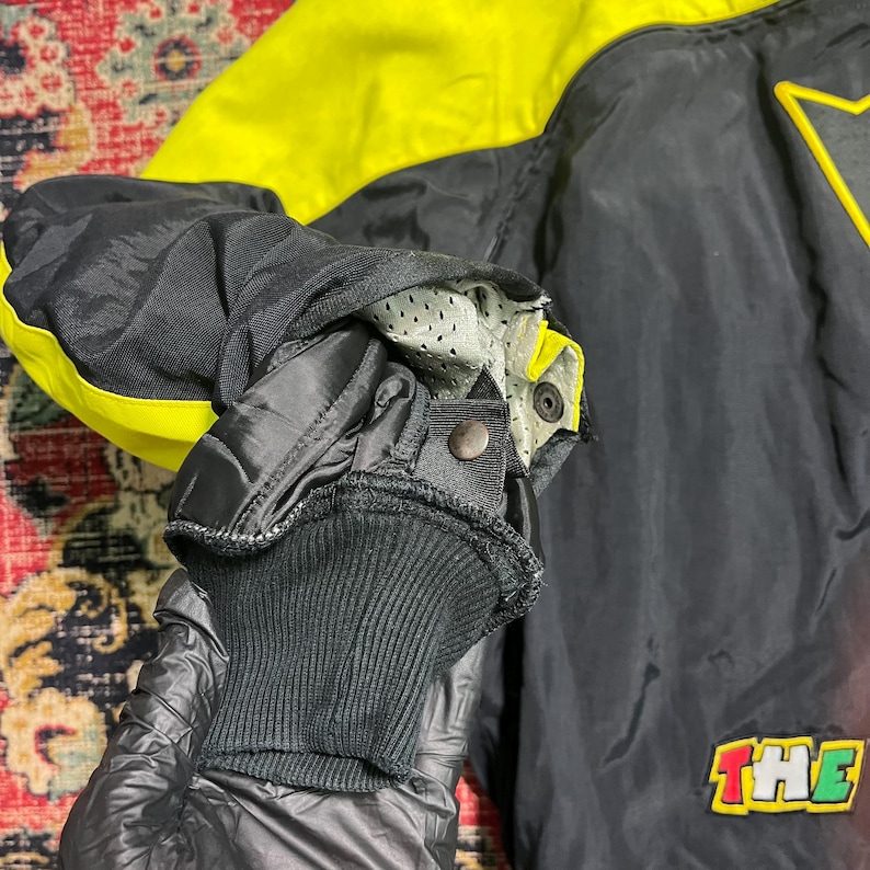 May include: Black and yellow jacket with a star design and the word "THE" in a colorful patch. The jacket has a ribbed cuff and a snap closure. The sleeve is black with yellow accents.