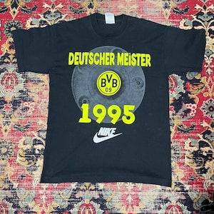 Könnte beinhalten: Schwarzes T-Shirt mit gelbem Schriftzug "DEUTSCHER MEISTER" über einem grauen Emblem mit "BVB 09" und dem Jahr "1995". Ein weißes Nike-Logo befindet sich darunter. Das Shirt hat kurze Ärmel.