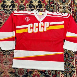 Peut inclure: Maillot de hockey rouge avec des détails blancs et jaunes. Le maillot présente les lettres "CCCP" en grandes lettres blanches sur la poitrine. Les manches ont des rayures blanches et jaunes. Un petit emblème est sur le haut de la poitrine.