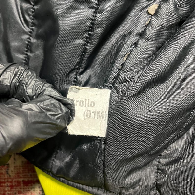 May include: Close-up of a black quilted jacket lining. A white tag with the text "Trollo (01M)" is visible. The jacket's interior is black, with visible stitching and a yellow underlayer.