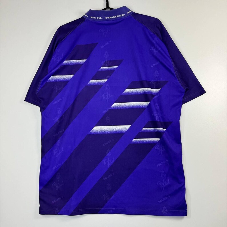 Puede incluir: Una camiseta de f&uacute;tbol vintage del Real Madrid en azul real, con rayas diagonales en azul oscuro y blanco. El cuello muestra "REAL MADRID" en blanco. La camiseta tiene mangas cortas y un dise&ntilde;o deportivo cl&aacute;sico.