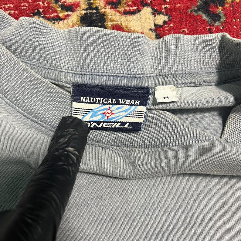 May include: A pale blue t-shirt with a crew neck. The label reads "NAUTICAL WEAR" above a blue and white design with the word "O'NEILL" below. The shirt is a medium size.
