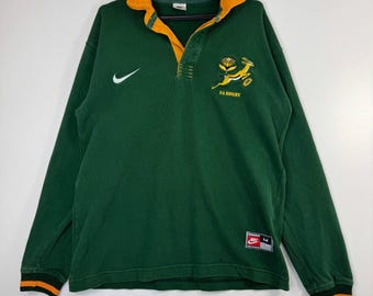 Puede incluir: Camiseta de rugby verde de manga larga con cuello y puños amarillos. Presenta un logotipo de Nike blanco y un logotipo con un saltamontes saltando y el texto "SA RUGBY". La camiseta tiene un cuello abotonado y una etiqueta Nike.