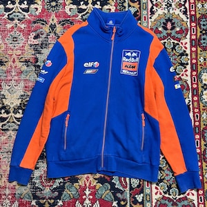 May include: Blue and orange zip-up jacket with the Red Bull KTM Tech3 logo. The jacket has orange panels on the sides and sleeves, with the Elf and WP logos. The jacket has a high collar and two front pockets with orange zippers.