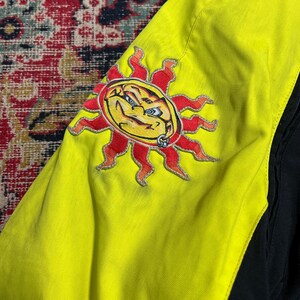 May include: A close-up of a yellow jacket sleeve with a stylized sun graphic. The sun has a yellow face, red rays, and a silver piercing. The jacket has black accents.