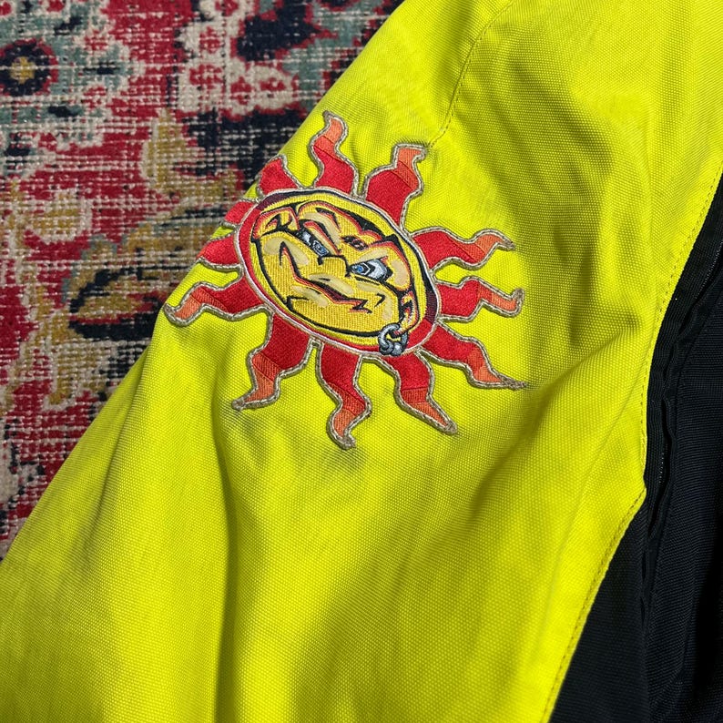 May include: A close-up of a yellow jacket sleeve with a stylized sun graphic. The sun has a yellow face, red rays, and a silver piercing. The jacket has black accents.