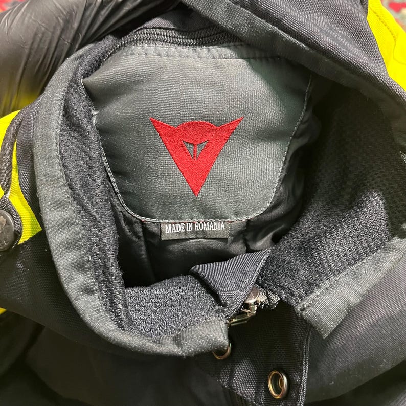 May include: Close-up of a black motorcycle jacket collar with a red logo and the text "MADE IN ROMANIA". The jacket has yellow accents and a zipper closure. The collar is lined with a soft, dark gray material.