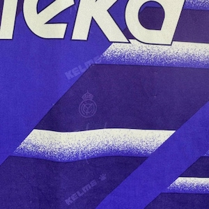 Puede incluir: Primer plano de una camiseta deportiva azul y blanca. La camiseta presenta la palabra "KEKO" en letras blancas grandes en la parte superior. La camiseta tiene un dise&ntilde;o geom&eacute;trico con la marca "KELME" y un escudo del equipo.