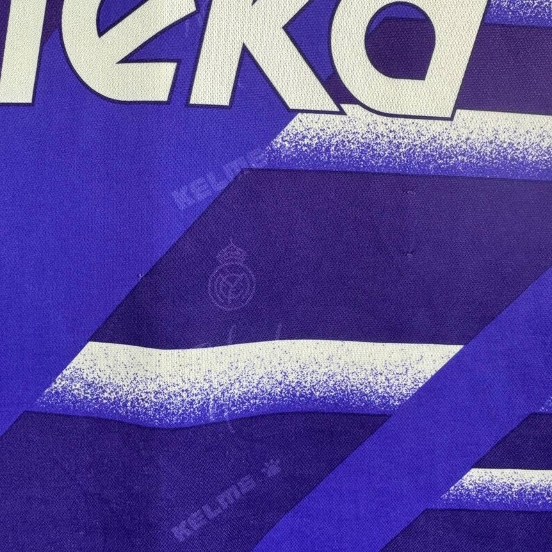 Puede incluir: Primer plano de una camiseta deportiva azul y blanca. La camiseta presenta la palabra "KEKO" en letras blancas grandes en la parte superior. La camiseta tiene un dise&ntilde;o geom&eacute;trico con la marca "KELME" y un escudo del equipo.