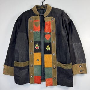Puede incluir: Chaqueta de cuero negro con paneles de pana y ante. La chaqueta presenta un diseño de patchwork colorido con apliques de ositos de peluche y botones decorativos. El cuello y los puños son de ante marrón.