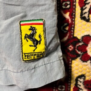 Puede incluir: Primer plano de una tela azul claro con un parche del logotipo de Ferrari. El parche presenta un caballo negro encabritado sobre un fondo amarillo, con la palabra "Ferrari" debajo. El parche tiene un borde verde, blanco y rojo.