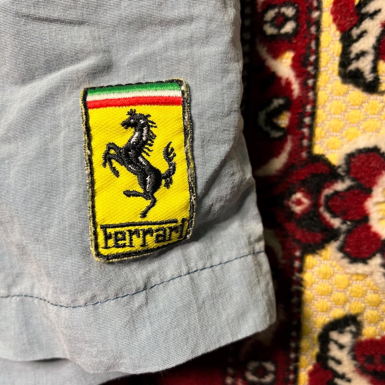 Puede incluir: Primer plano de una tela azul claro con un parche del logotipo de Ferrari. El parche presenta un caballo negro encabritado sobre un fondo amarillo, con la palabra "Ferrari" debajo. El parche tiene un borde verde, blanco y rojo.