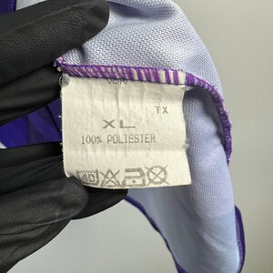 Puede incluir: Primer plano de una etiqueta de ropa con la talla "XL" y la composici&oacute;n "100% POLIESTER". La etiqueta blanca con texto negro est&aacute; cosida a una prenda morada y azul claro de tejido texturizado.