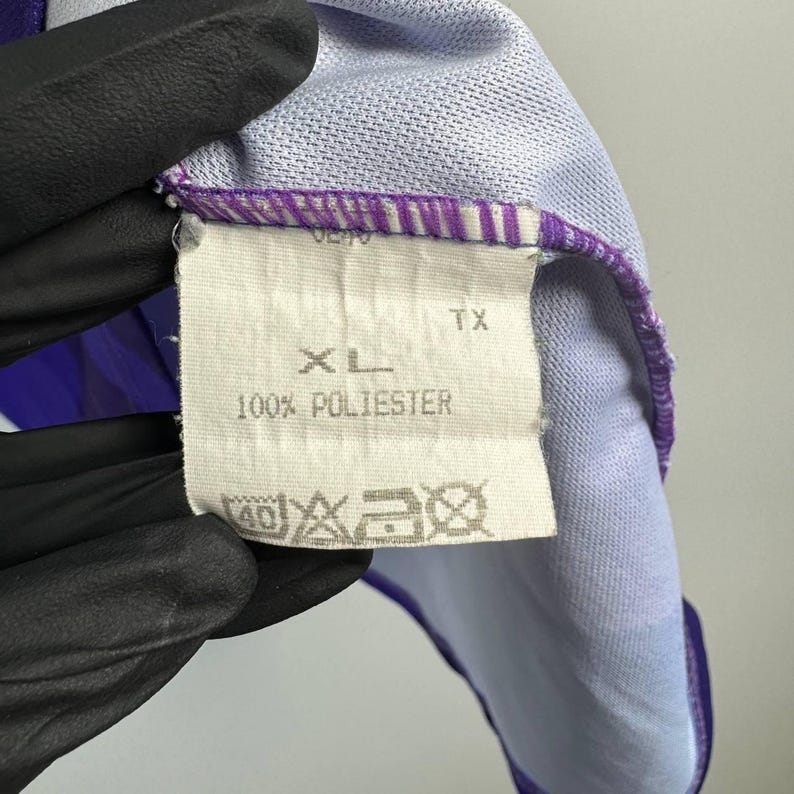 Puede incluir: Primer plano de una etiqueta de ropa con la talla "XL" y la composici&oacute;n "100% POLIESTER". La etiqueta blanca con texto negro est&aacute; cosida a una prenda morada y azul claro de tejido texturizado.