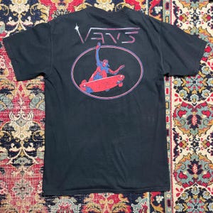 以下が含まれることがあります： 黒いTシャツ。ピンクと青の楕円の中に赤いスケートボードに乗ったスケーターのグラフィック。 「VANS」の文字が楕円の上にピンクと白で書かれています。 このシャツは半袖です。