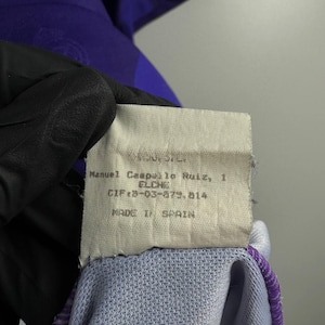 Puede incluir: Primer plano de una etiqueta con texto. La etiqueta es blanca con texto negro que dice "Manuel Campallo Ruiz, 1 ELCHE CIF:8-03-879.814 MADE IN SPAIN". La etiqueta est&aacute; adherida a una prenda morada y azul.