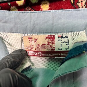 Puede incluir: Primer plano de una chaqueta Fila azul claro con una etiqueta Ferrari. La etiqueta presenta una ilustraci&oacute;n vintage y el texto "OFFICIAL LICENSED PRODUCT". La chaqueta tiene un forro de malla y una etiqueta de talla peque&ntilde;a.