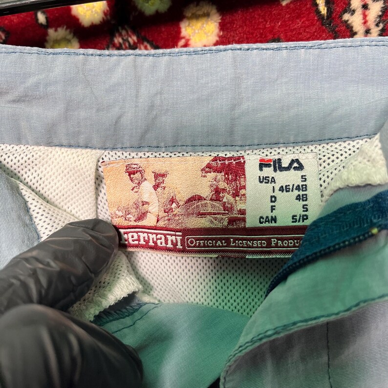 Puede incluir: Primer plano de una chaqueta Fila azul claro con una etiqueta Ferrari. La etiqueta presenta una ilustraci&oacute;n vintage y el texto "OFFICIAL LICENSED PRODUCT". La chaqueta tiene un forro de malla y una etiqueta de talla peque&ntilde;a.