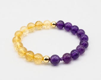 May include: A beaded bracelet featuring a sequence of translucent yellow citrine beads and deep purple amethyst beads, separated by two polished gold-tone metal spheres. The beads are arranged in a circular pattern on a plain white background.