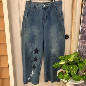 May include: High-waisted blue denim jeans featuring a wide-leg silhouette and curved side seams. The front of the right leg is decorated with a large denim star patch and a vertical row of smaller stars in varying shades of blue and white.
