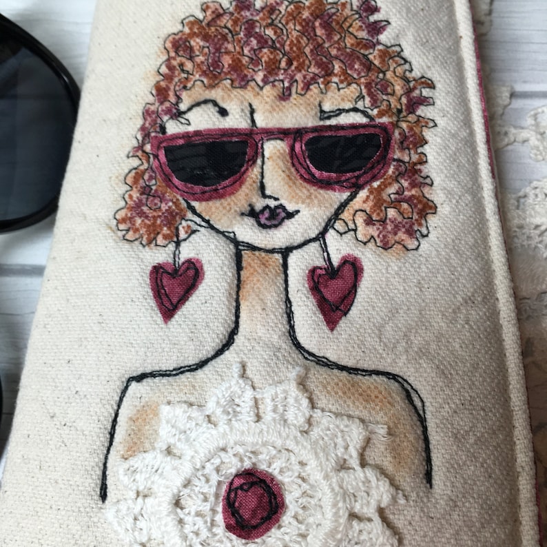 Op de afbeelding: Een witte canvas tas met een geborduurde afbeelding van een vrouw met krullend bruin haar die een roze zonnebril en hartvormige oorbellen draagt. De vrouw draagt een wit gehaakt topje met een roze bloem in het midden.