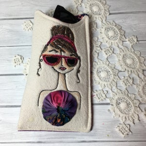 Op de afbeelding: Een wit stoffen brilletje met een kleurrijk geborduurd ontwerp van een vrouw met een zonnebril en een bloem. Het etui heeft een paarse voering.