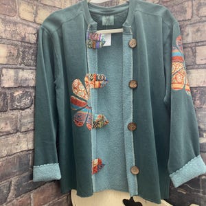 May include: A teal long-sleeve cardigan featuring a row of four brown wooden buttons, a light blue inner trim, and decorative fabric patches with colorful floral patterns on the front and sleeve. The cuffs have a scalloped edge finish.