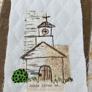 May include: A quilted fabric art piece featuring a church illustration. The church has a steeple with a cross, a clock, and a door. The text "Jesus loves me" is stitched below the church. The background is white with a diamond pattern.