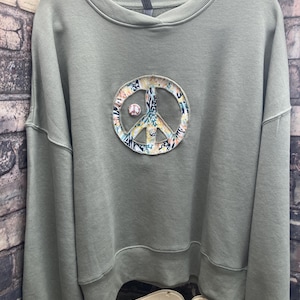 May include: An olive green long-sleeve sweatshirt featuring a large circular peace sign appliqué on the chest. The peace sign is filled with a multi-colored patterned fabric in shades of orange, blue, and black. A small circular pin is attached.