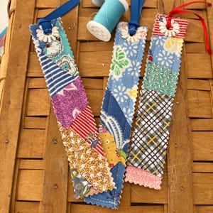 May include: Three rectangular fabric bookmarks featuring patchwork designs with various floral, striped, plaid, and geometric patterns in blue, red, yellow, and white. Each bookmark has pinked edges, a white crocheted ring at the top, and a ribbon tie.