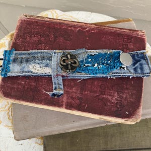 May include: A stack of vintage books with a denim and turquoise embellished book wrap. The wrap features a metal clasp and a silver button with the number 1966. The books have burgundy, gray, and olive green covers.