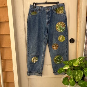 May include: Blue denim jeans featuring a high-waisted fit with a button and zipper fly closure. The jeans are decorated with multiple circular, hand-painted green and yellow designs, including a peace sign on the lower left leg and cuffs.